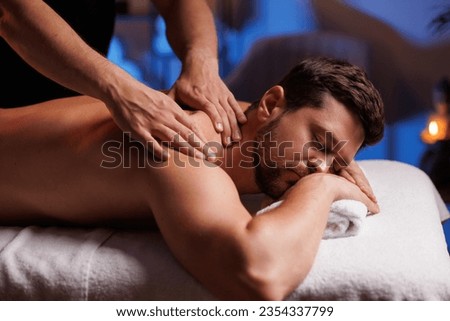 Handsome man having restorative back massage in spa salon, enjoying relaxing atmosphere, recharging after work. Masseuse gives therapeutic back massage to a visitor, the concept of healthy lifestyle. Royalty-Free Stock Photo #2354337799
