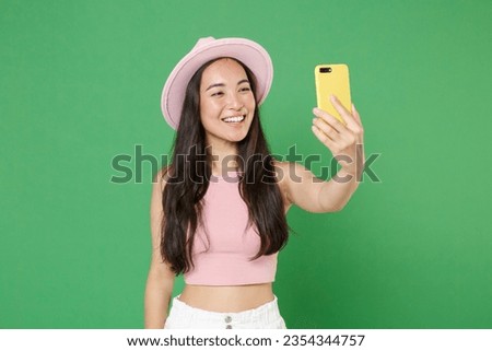 Smiling woman of Asian ethnicity wear casual pink clothes posing isolated on green wall background studio portrait. People lifestyle concept. Mock up copy space. Doing selfie shot on mobile cell phone
