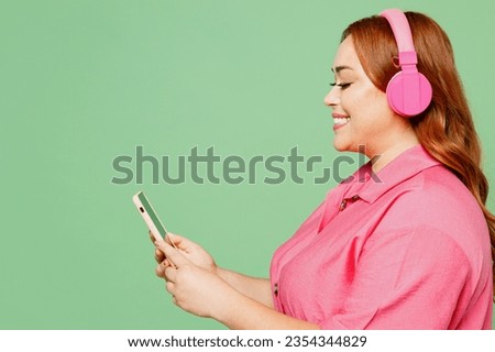 Side view young chubby overweight redhead woman wear casual clothes pink dress listen music in headphones use mobile cell phone isolated on plain pastel light green color background. Lifesyle concept