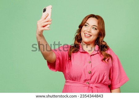 Young chubby overweight redhead woman wear casual clothes pink dress doing selfie shot on mobile cell phone post photo on social network isolated on plain light green color background Lifesyle concept