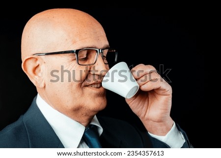 aged bald man is captured up-close, relishing genuine Italian espresso from a suitably-sized cup. Dressed in a dark jacket and white shirt, with black glasses on, he's a picture of contentment. Note:  Royalty-Free Stock Photo #2354357613