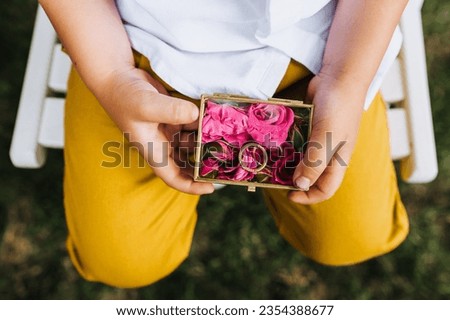 A child, a boy, holds in his hands a glass box with flowers, golden rings with words of love forever at the wedding ceremony. Photo close-up, portrait.