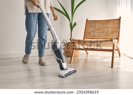 Side view of crop faceless housewife in casual outfit and slippers cleaning laminate floor with modern upright vacuum cleaner in minimalist light apartment Royalty-Free Stock Photo #2354398619