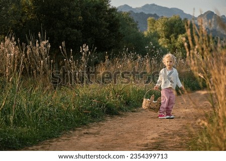 Full body adorable curious girl with wavy hair standing with wicker basket on narrow dirt road and looking at camera while spending warm summer day in peaceful nature Royalty-Free Stock Photo #2354399713