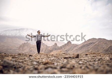 Full length ground level of Hispanic male tourist standing with outstretched arms on dry rocky ground against mountain Teide and looking away in Tenerife Canary Islands Spain Royalty-Free Stock Photo #2354401915
