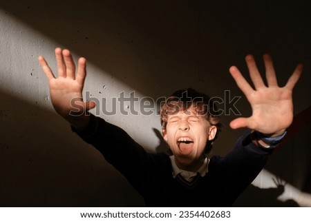 Excited preteen boy standing alone in dark room shouting and showing stop gesture with palms of hands while reacting and resisting sudden bright sunlight