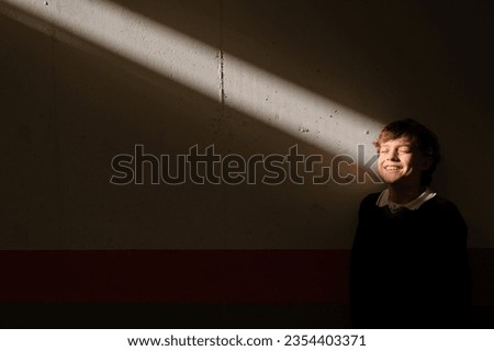 Positive cute preteen boy standing with closed eyes at wall in dark room catching sunbeam in face Royalty-Free Stock Photo #2354403371