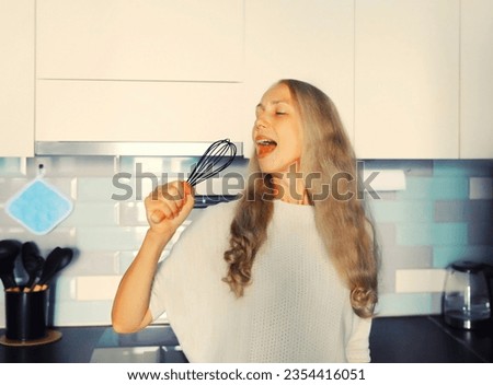 Happy cheerful middle-aged gray-haired woman singing with whisk having fun in the kitchen at home