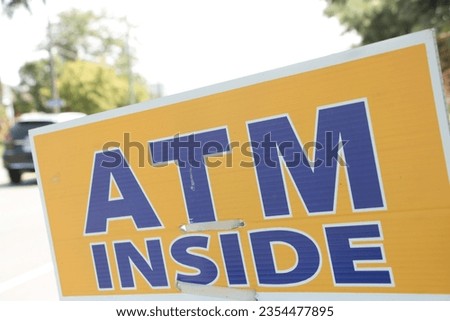 atm inside sign in black writing on yellow background with bright background behind it with car in summer