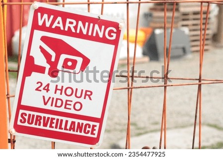 warning 24 hour video surveillance sign on orange locked gate for dirt yard parking lot with containers bins skids palettes, close up looking down, red and white