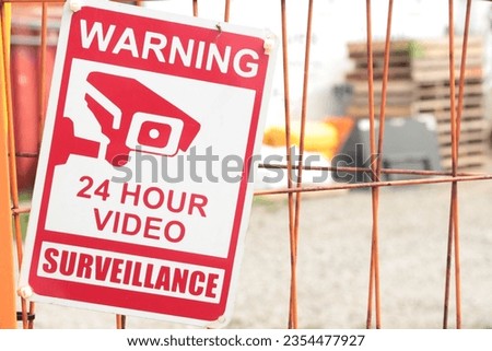warning 24 hour video surveillance sign on orange locked gate for dirt yard parking lot with containers bins skids palettes, close up, red and white