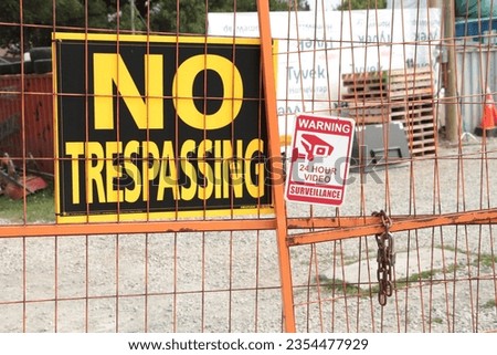 no trespassing warning 24 hour video surveillance signs on orange locked gate for dirt yard parking lot with containers bins skids palettes, close up, yellow black red and white