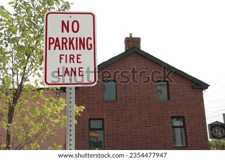 no parking fire lane vertical rectangle vintage style sign on post with red brick house home building behind tree and sky, red writing caption text on white background