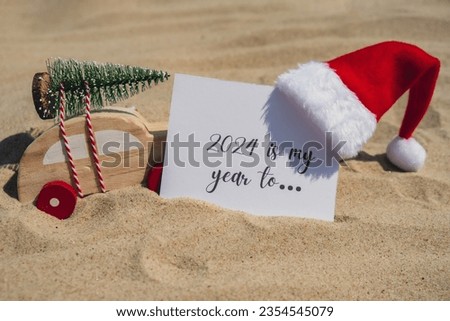 2024 IS MY YEAR TO.. text on paper greeting card on background sandy beach sun coast. Christmas balls Santa hat New Year New Me Resolutions decoration. Summer vacation decor. Holiday concept calendar