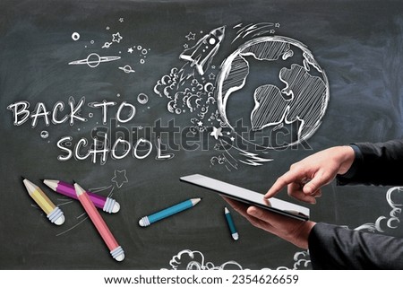 Close up of businessman hand holding and pointing at tablet with abstract back to school sketch with pencils and rocket on chalkboard wall background. Education and knowledge concept