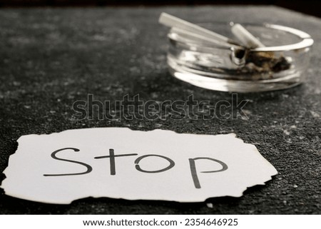 Ashtray with burnt cigarettes and word Stop written on paper on black textured table, closeup. No smoking concept