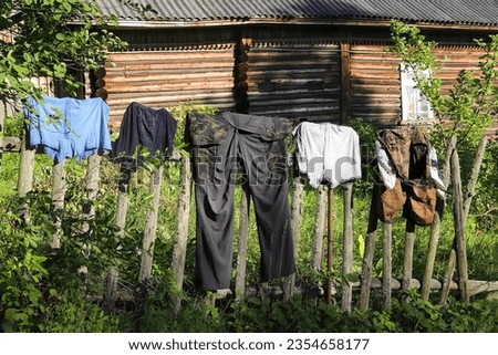 Washed men's work clothes are dried after washing on a dilapidated mossy fence in the village on a clear sunny day. In the background is a log Russian house. Romance of the Russian hinterland
