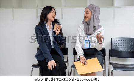 Two friendly and happy young Asian female job candidates enjoy talking while waiting in front of the room for a job interview. First jobbers, internship, employment, job hiring Royalty-Free Stock Photo #2354661483