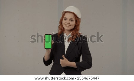 Architect woman showing smartphone green screen and thumb up. Portrait of a business woman in a helmet and a smartphone in her hands on a gray background in the studio. Advertising area, workspace.