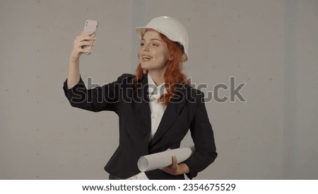 Woman architect talking on video call, taking selfie using smart phone. A woman in a helmet and with blueprints in a studio on a gray background.