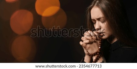 Praying young woman with rosary beads on dark background with space for text