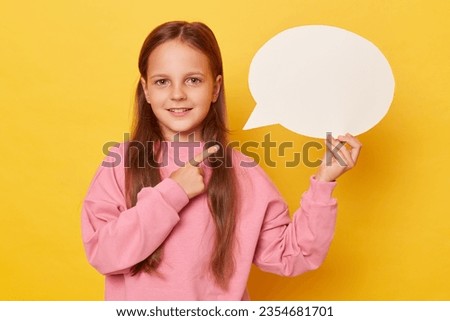 Cute smiling happy little girl with long hair holding pointing finger at empty speech bubble wearing pink sweatshirt isolated over yellow background looking at camera