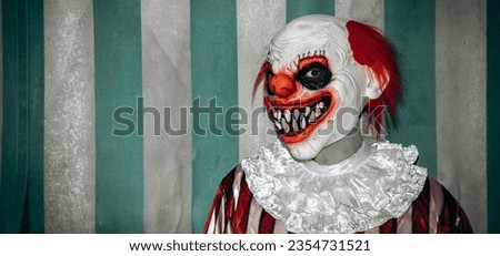 a mad evil redhead clown, wearing a white and red striped costume with a white ruff, stands with a creepy smile in front of the weathered tent of the circus, in a panoramic format to use as web banner