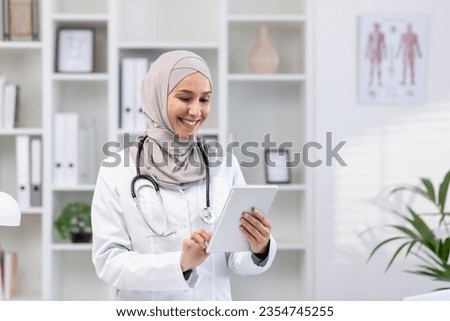 Young beautiful female doctor in hijab using tablet computer, doctor worker in white medical coat and stethoscope inside clinic office at workplace, smiling muslim woman using online application.