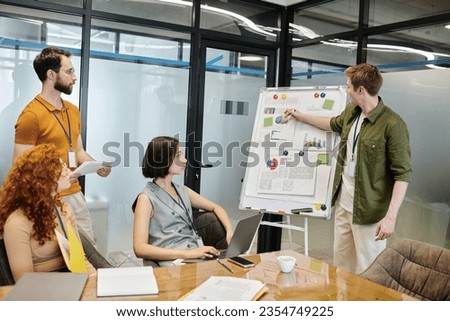 young manager pointing at graphs on flip chart near creative team in meeting room, startup project Royalty-Free Stock Photo #2354749225