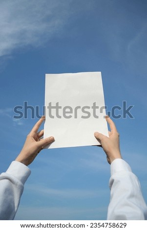 Hands holding up a blank sheet of paper outdoors, on a blue sky background.