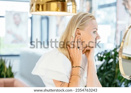 Beautiful middle age blond woman choosing earings looking at mirror in a modern jewelry store. Luxury jewelry store concept. Jewelry boutique.