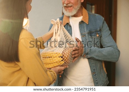 Helping neighbours. Young woman with net bag of products visiting senior man outdoors on sunny day, closeup Royalty-Free Stock Photo #2354791951