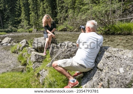 Mature photographer man taking photos of a blonde middle-aged model near a stream in the Italian Dolomites