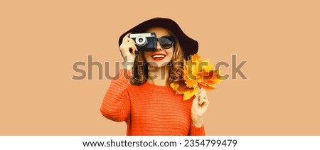 Autumn color style outfit, stylish young woman photographer with film camera and yellow maple leaves wearing sweater and hat on brown background
