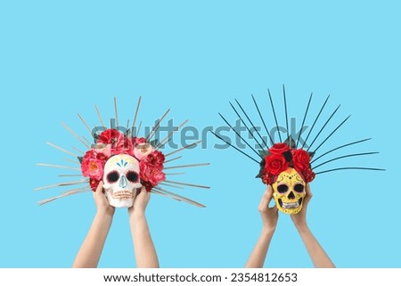 Female hands holding painted human skulls for Mexico's Day of the Dead (El Dia de Muertos) with flowers on blue background