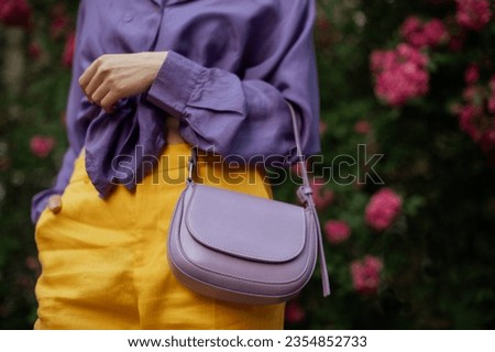 Pale purple faux leather trendy small bag, handbag in fashionable woman`s hand. Model wearing violet blouse, yellow trousers. Copy, empty space for text Royalty-Free Stock Photo #2354852733