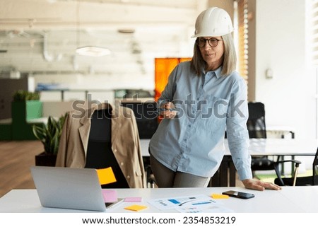 Portrait of confident serious senior woman, architect, engineer using laptop working project looking at computer monitor standing in modern office. Successful business concept