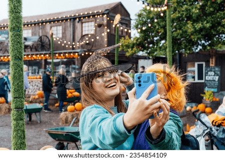 Two laughing female friends in fun glasses and witch hat having fun and making selfie on phone on decorated pumpkin farm. Selecting Thanksgiving and Halloween holidays decor. Autumn fall festive mood.