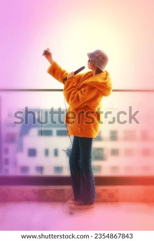 Teenage girl taking a selfie or streaming a live video while standing by the glass railing of a balcony at an observation deck overlooking the city.