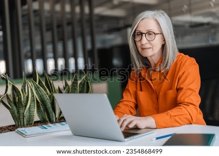 Confident serious mature businesswoman wearing stylish eyeglasses, orange shirt using laptop working online in modern office. Manager planning project, typing on keyboard. Successful business, career