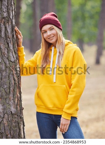 Attractive blue eyed blonde woman walk on the park. Girl wear yellow hoodie and burgundy hat, look happy and smiles. Portrait of a joyful young woman enjoying in autumn park.