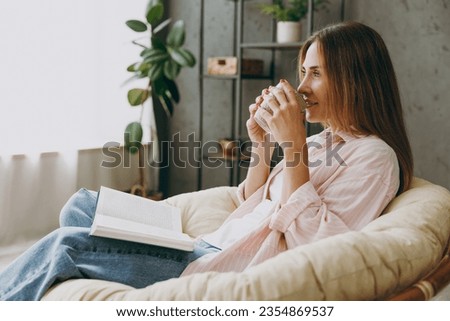Side profile view young woman wear casual clothes sits in armchair read book drink coffee tea stay at home hotel flat rest relax spend free spare time in living room indoor Lifestyle lounge concept Royalty-Free Stock Photo #2354869537
