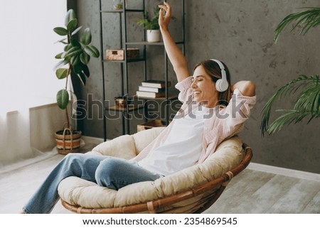 Side view young cool woman wear casual clothes sits in armchair listen to music in headphones stay at home hotel flat rest relax spend free spare time in living room indoor. Lifestyle lounge concept Royalty-Free Stock Photo #2354869545