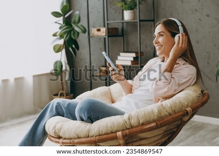 Young woman wears casual clothes sits in armchair listen to music in headphones use mobile cell phone stay at home hotel flat rest spend free spare time in living room indoor. Lifestyle lounge concept Royalty-Free Stock Photo #2354869547