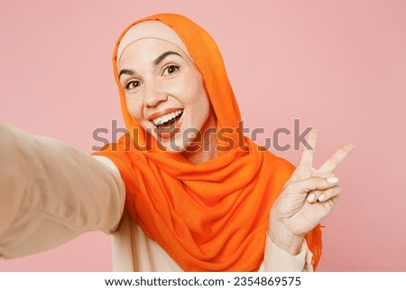 Young arabian asian muslim woman wears orange abaya hijab doing selfie shot pov on mobile cell phone isolated on plain light pink background studio portrait. Uae middle eastern islam religious concept
