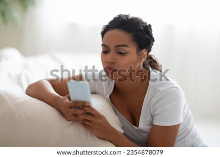 Upset Yong Black Woman Reading Message On Smartphone At Home, Bored Millennial African American Female Browsing Social Networks On Mobile Phone While Relaxing On Floor In Bedroom, Closeup Royalty-Free Stock Photo #2354878079