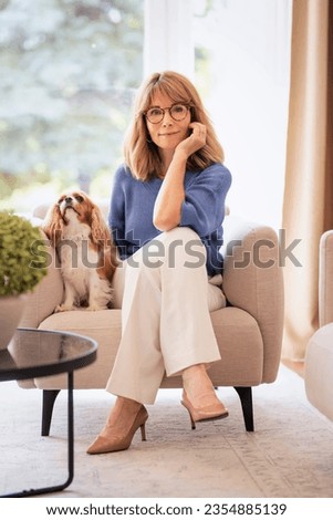 Full length of an attractive blond haired woman relaxing in armchair at home with her cute puppy. Mid aged female looking at camera and cheerful smiling. Woman wearing blue shirt and white pants