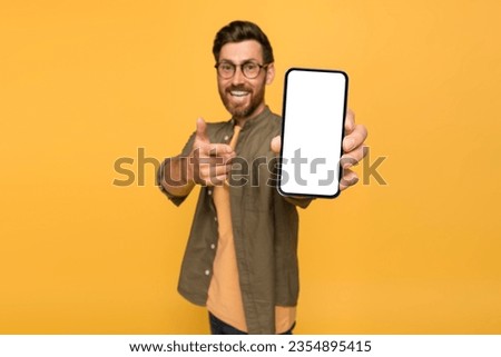 Positive european middle aged man pointing at smartphone with white blank screen in his hand and smiling, isolated on yellow studio background, showing new online offer, mockup