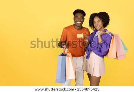 Happy millennial black guy and lady with many bags show credit card, recommend shopping, isolated on yellow studio background. Sale, finance, credit recommendation, ad and offer