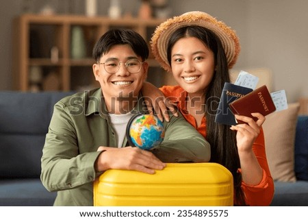 Portrait of smiling japanese tourists couple holding travel tickets and passports, posing with suitcase and world globe at home, smiling to camera. Young globetrotters going on honeymoon vacation Royalty-Free Stock Photo #2354895575